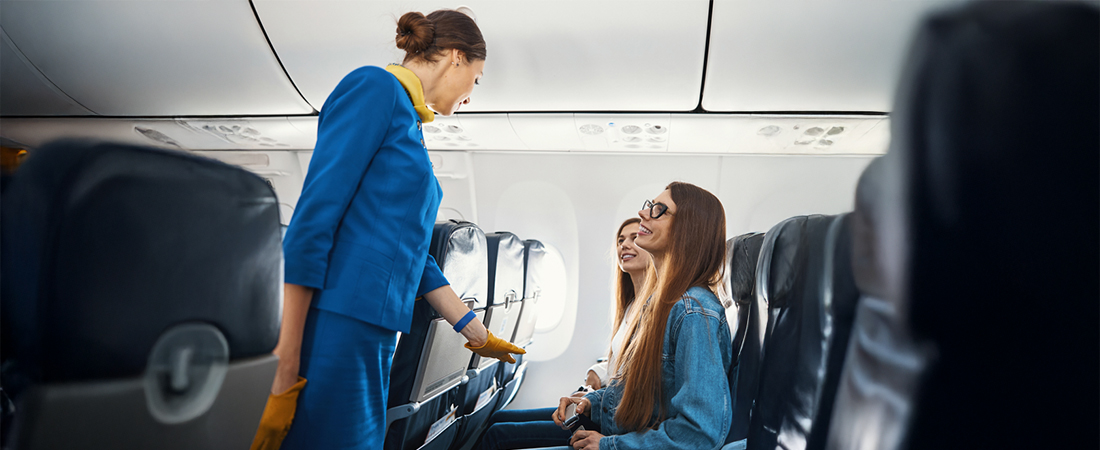 Una azafata de vuelo interactúa de manera amable y profesional con una pasajera sentada en la cabina del avión.