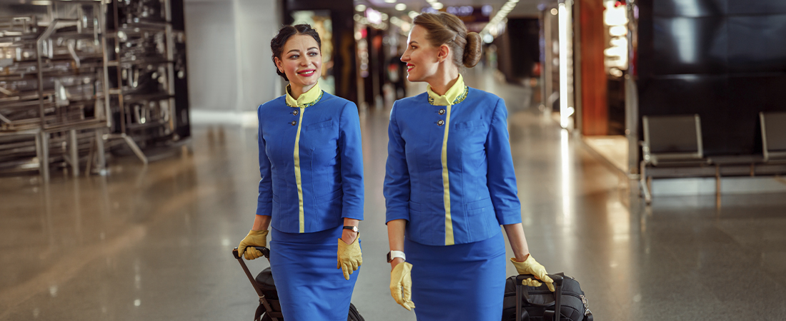 Dos azafatas de vuelo con uniforme azul brillante caminando juntas por la terminal de un aeropuerto, tirando de sus maletas de mano.